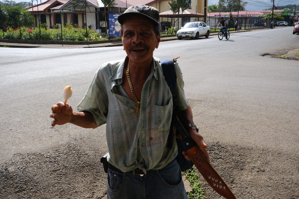 Portrait d'un homme moustachu âgé souriant, portant une casquette, une chaîne en or, tenant une glace et une machette dans son étui