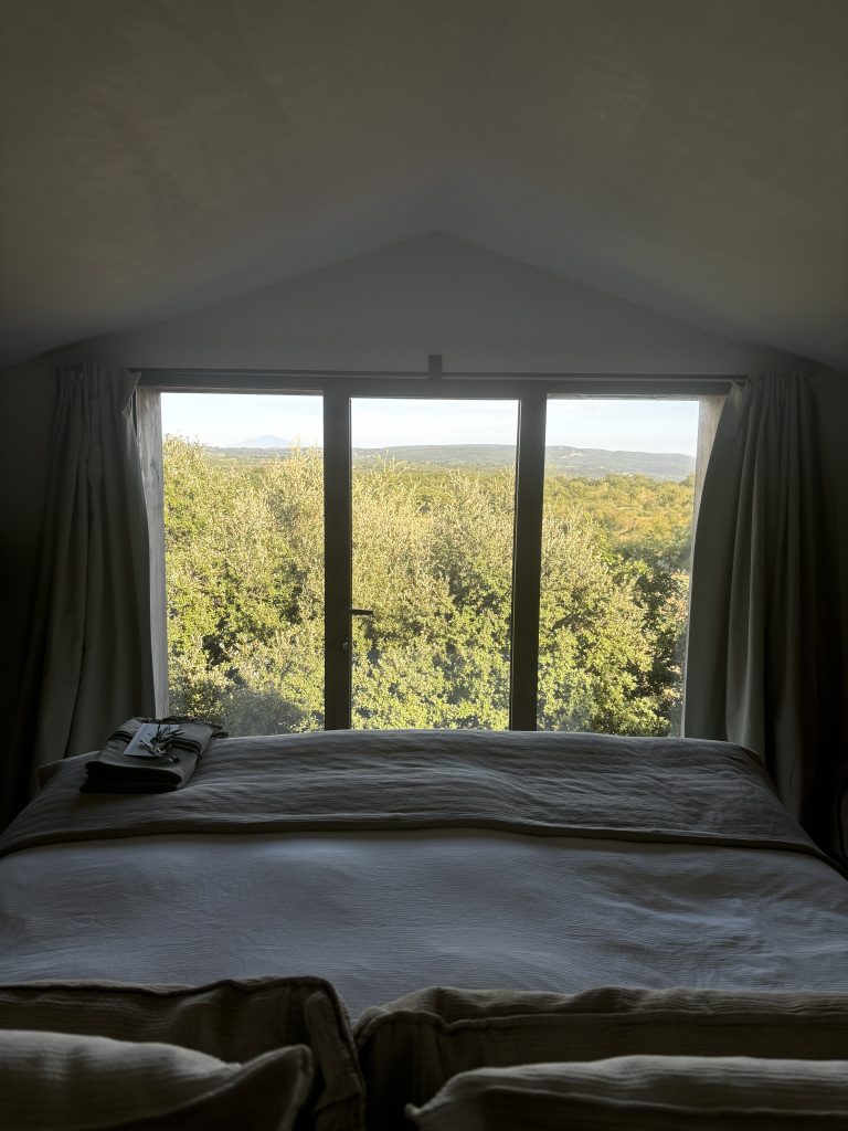 Vue depuis un lit blanc vers une grande fenêtre encadrée de rideaux clairs, révélant une forêt dense sous un ciel lumineux.