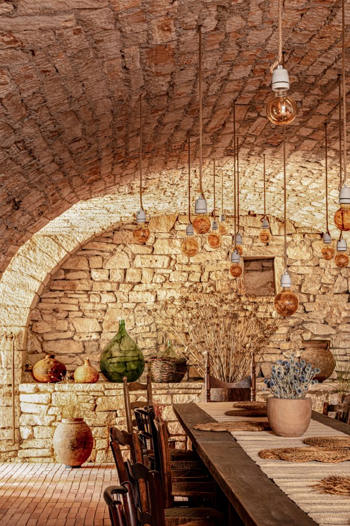 Salle à manger rustique sous un plafond voûté en pierre apparente, avec une longue table en bois sombre éclairée par de nombreuses ampoules suspendues à des fils.