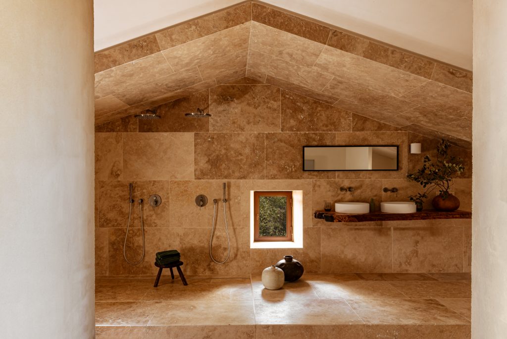 Salle de douche luxueuse entièrement revêtue de pierre beige, comprenant deux douches de tête, une petite fenêtre carrée donnant sur la verdure et une étagère en bois brut avec deux vasques.